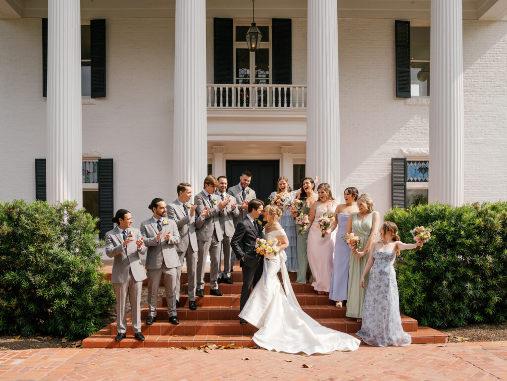 Newlyweds kissing on the front steps of Woodbine Mansion as their wedding party celebrates around them.
